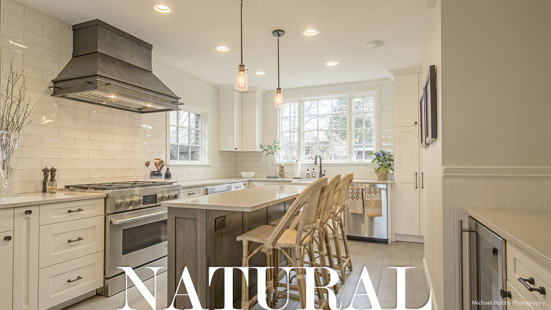 bright natural light kitchen with white cabinets and subway tile covering all walls, angular metal steel range hood in dark washed patina is alone on wall above range, two Edison bulbs above modest small island that has three barstools