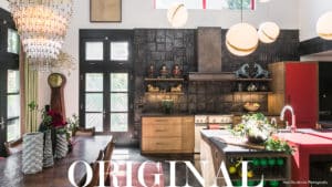 Raw Urth modern custom metal range hood in a highly decorated kitchen with modern light fixtures, ornaments on shelves, and wooden cabinets; with the word "original" written below