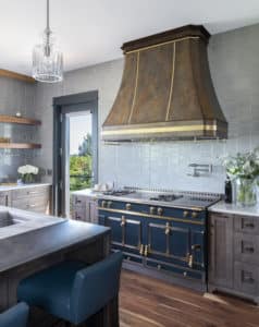 Grand range hood that resembles a rusted metal because of patina and coloring with brown, gray, red tones. Burnished Brass band around the apron, two channel seams down the face, and a toolbar hanging on the underside of three sides. Style is Mary Jane by Raw Urth Designs.