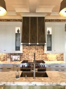 Patina'd in a dark wash finish, this metal range hood slowly slopes out from high decorated white trimmed ceiling. Red brick backsplash goes under and above cabinets. Hood surrounded by white cabinets with thick and elegant crown molding.