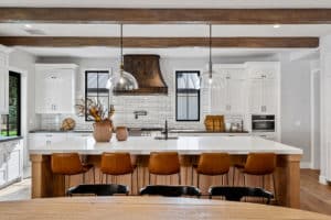 Centered in kitchen surrounded by white tile and cabinets with windows on either side of hood. Finished in a Reclaimed Steel Patina built by raw Urth Designs. Style is a custom winter park design. Camel colored leather barstools sit across from large island and range hood.