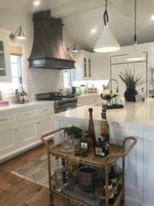 Large custom antique zinc hood straddles beam in high ceiling white cabinet kitchen. In forefront, wicker bar cart with alcoholic drinks on display. Two white cone pendant lamps hang above spacious island.