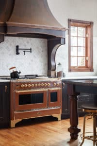Cherry hills metal range hood in a rustic kitchen over a gas powered brown stove and dark colored cabinets