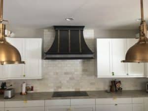 Modern yet traditional style custom metal range hood. Body of the hood slopes out on either side and finished in antique charcoal gray steel. Three straps on the face are an Antique Brass patina. Industrial Brass light fixtures hang in the forefront above the island.