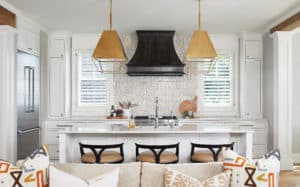 Dark curved metal range hood centered on wall between two windows. Behind is a small pattern tile backsplash. Two Brass pendant lights hang above the island. In the forefront, the top of a neutral sofa with colorful pillows shows sneakily.
