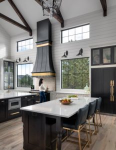 custom metal range hood in a kitchen with a high vaulted ceiling flanked by two oversized kitchen windows. Range hood is Black with Brass details on a white wall. Other items in kitchen include white marble counters and black cabinets for a tuxedo kitchen look.