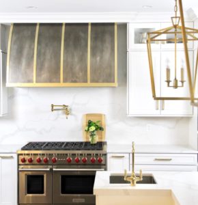 centered between white cabinets with brass hardware, cool toned brown light washed patina hood body with burnished brass straps and bands for design detail. matches light fixture, sink faucet, and Wolf cook top.