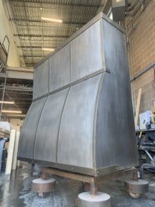 range hood in work shop. steel curved outward front face and tall chimney. light washed steel patina with foundry steel straps to look like brass or bronze.
