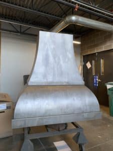 custom bonner peak range hood fabricated out of steel and patina'd to be light washed. bottom portion of hood billows out, comes inward, and slopes in for the chimney cover piece.