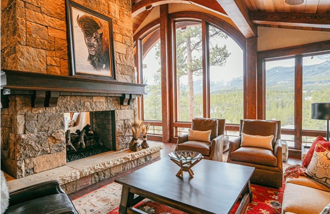 A cozy living room is enhanced by a stone fireplace with bison artwork above it, large windows showcasing pine trees and mountain views, complemented by a wooden coffee table, brown armchairs, and patterned rug for a warm rustic feel. Featured on blog-mountainliving.