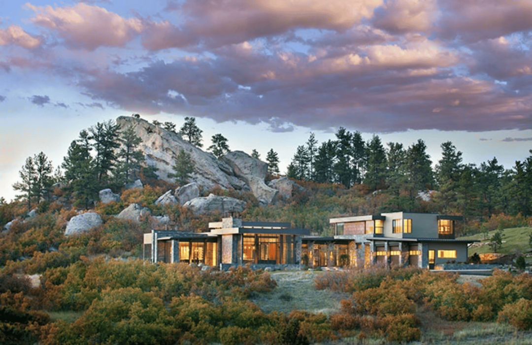 Amidst a rocky landscape dotted with trees and shrubs, 'blog-coloradohome' stands as a modern house with large windows. It contrasts beautifully with the natural scenery as it shines from within under a dramatic sky scattered with clouds.