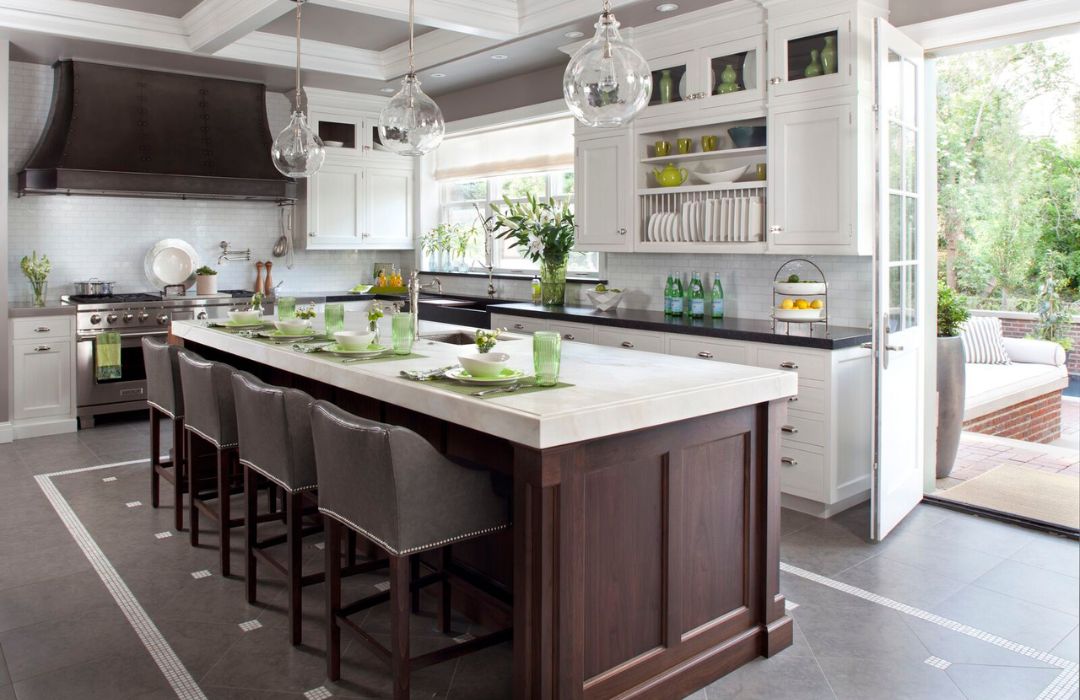 A modern kitchen showcases a large island with a white countertop and wooden base, paired with five gray chairs and green-white place settings. The space features white cabinets, stainless steel appliances, and a glass door leading outside. Product source: blog-luxeinteriors.
