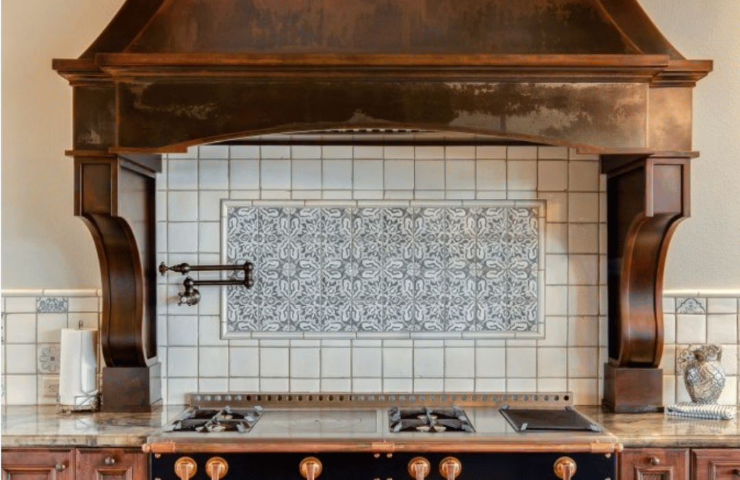 The rustic kitchen showcases the blog-steeligtheshow, featuring a large ornate wooden range hood above the stove. Intricately patterned tiles form the backsplash, while vintage-style knobs and burners adorn the stovetop. Warm, earthy tones complete the design.