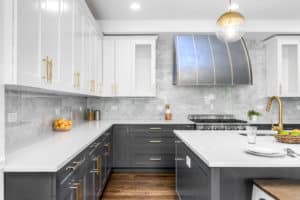 Gray and blue toned Metal range hood, concave front face, with brass ribs and rivets. Dark grey base cabinets and white wall cabinets have brass pulls and handles. Marbled subway tile backsplash surrounds hood to high ceiling. Light, airy and fresh modern space.