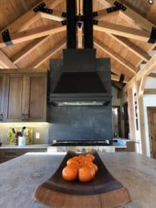 Decorative metal black range hood installed within a lodge kitchen. Hood has toolbar on apron. The sides angle in and extend up to a high vaulted ceiling with large wooden beams, clad with dark metal brackets for support. Behind the hood, is a gray and blue patina tile backsplash. Cabinets are a walnut simple door frame, with under cabinet lighting that glows down onto the quartz countertop. A bowl of small oranges centered on the island sits in the forefront.