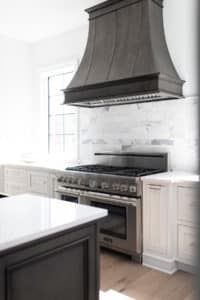 decorative brown toned gray traditional steel range hood by raw urth designs. elegant trim on top and bottom edge, with three straps on hood body. sits on wall in bright, light colored kitchen. below hood is marble subway tile backsplash and a wide Thermador cook top oven range.