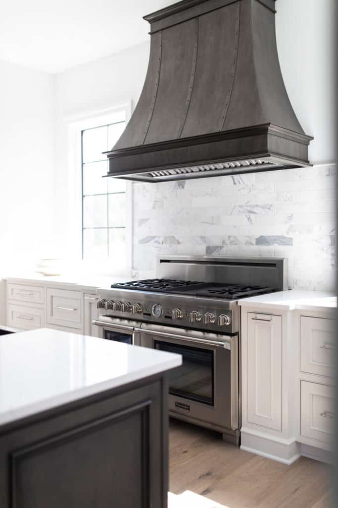 An example showing a decorative metal range hood between two kitchen windows with range hood by Raw Urth Designs.