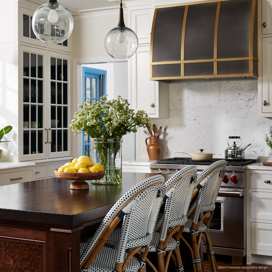 Aspen gallery1✅ (3) The Aspen gallery1✅ (3) features a stylish kitchen with white cabinets, a dark island countertop, woven bar stools, pendant lights, a large black-and-gold range hood over the stove, plus floral accents and a bowl of lemons.