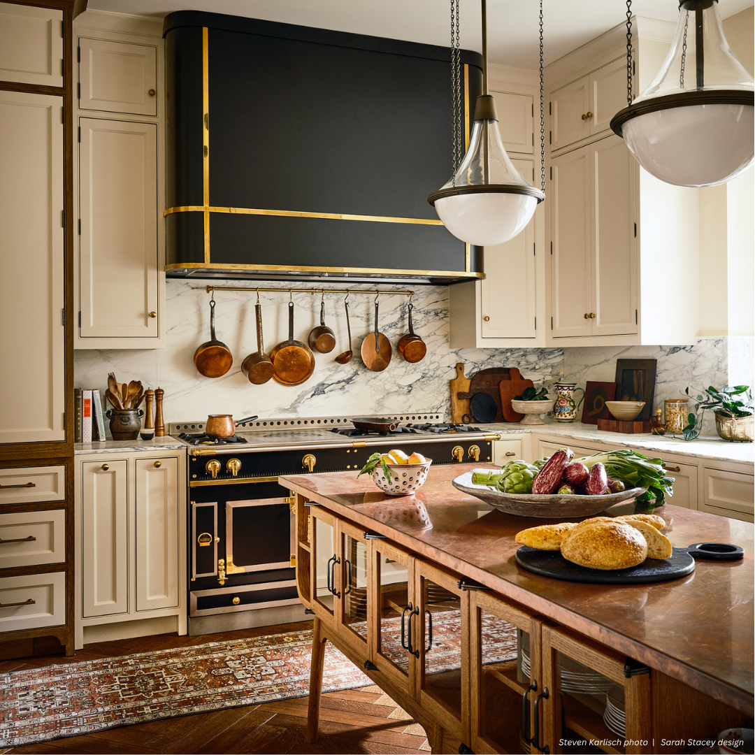 The Broadmoor gallery2 features a bright kitchen with white cabinets, a black and gold stove, copper pans on the wall, marble backsplash, pendant lights, and a wooden island topped with fresh vegetables and bread.