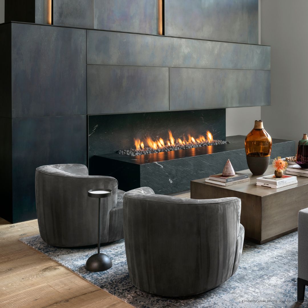 Cladding Gallery 1 The modern living room features a sleek horizontal fireplace set in smooth, dark Cladding Gallery 1 panels, with two curved gray chairs facing the fire on a patterned rug. A wooden coffee table with decorative vases and books completes the cozy, stylish ambiance.
