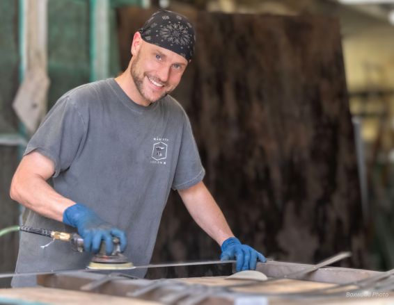 In a workshop, a person in a gray T-shirt, bandana, and gloves is smiling while using the Darin Bio sanding tool on wood, surrounded by wooden materials.