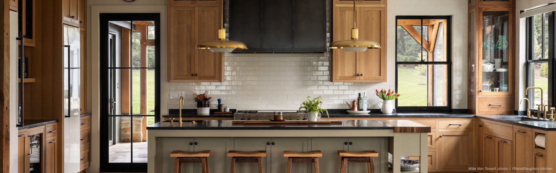 The spacious kitchen showcases the EVERGREEN Aspen Classic: a large island with four wooden stools. Light wood cabinets contrast with black countertops, while two pendant lights hang above. Large windows offer views of the greenery outside.