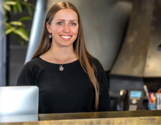 A woman with long brown hair, wearing a black top and necklace, stands smiling behind a counter. The Ella Bio-3 laptop and an office phone can be seen in the background.