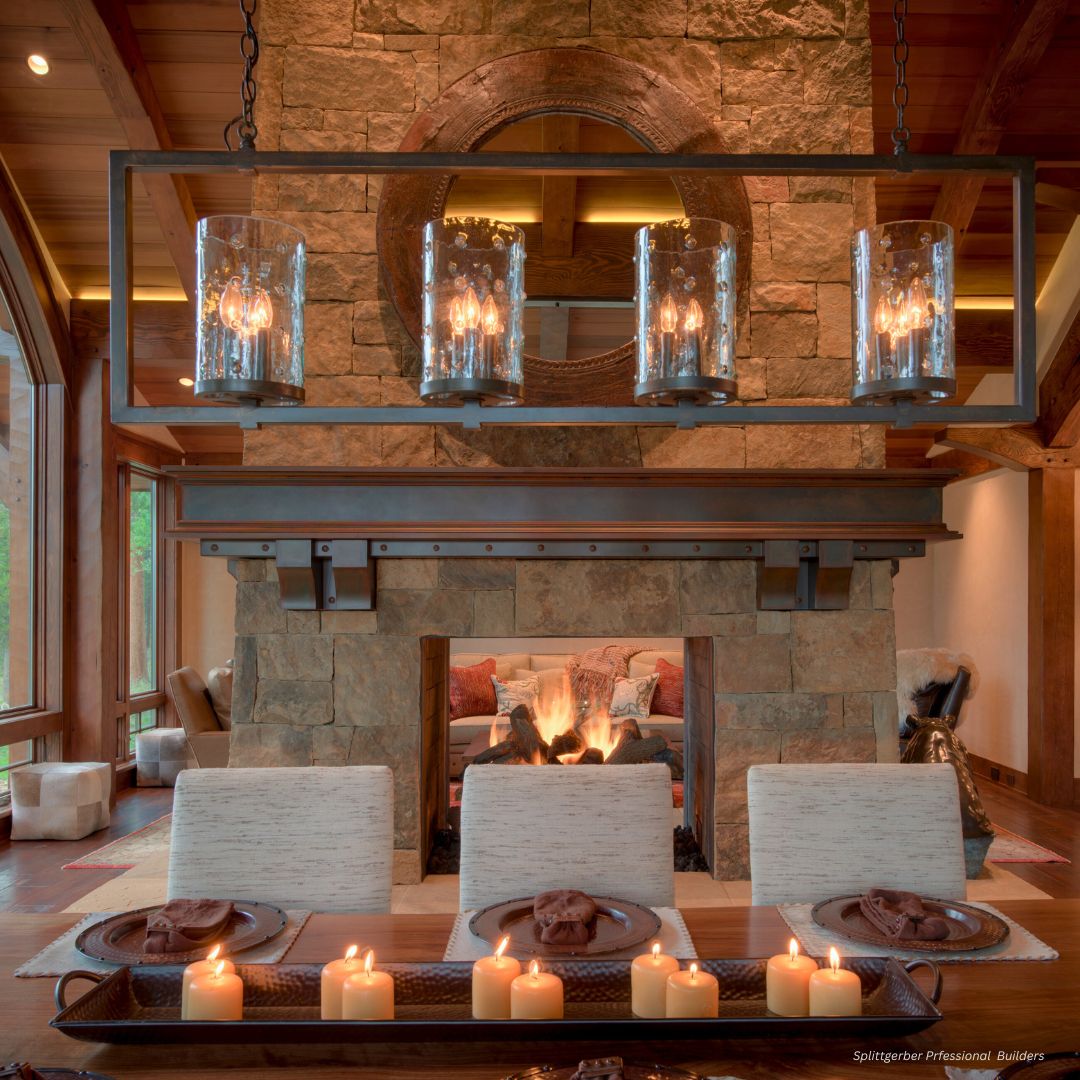 FP Mantel 1 A cozy living room features a rustic FP Mantel 1 stone fireplace and wooden ceiling. The dining table, adorned with plates, napkins, and a row of lit candles, sits beneath a large rectangular chandelier with glass lights.