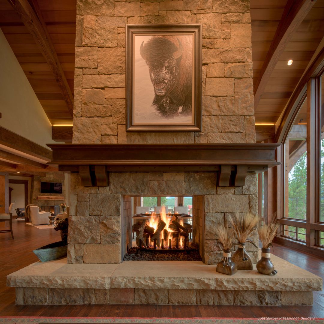 FP Mantel 2 A stone double-sided fireplace (FP Mantel 2) with a roaring fire and bison picture above, surrounded by wooden beams and large windows. Two vases filled with wheat are on one side of the mantel, creating a cozy rustic ambiance.