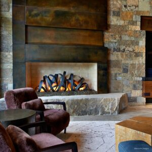 Modern living room with the FP Sculpture 2, a large metal and stone fireplace. The space includes plush brown armchairs and a wooden table set on a light patterned rug, creating a cozy atmosphere.