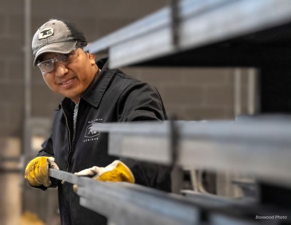 Wearing safety glasses and a cap, a person smiles while pulling steel beams in an industrial setting. They wear a dark logo jacket labeled "Gustavo bio" with yellow gloves, against a background of blurred metal beams.