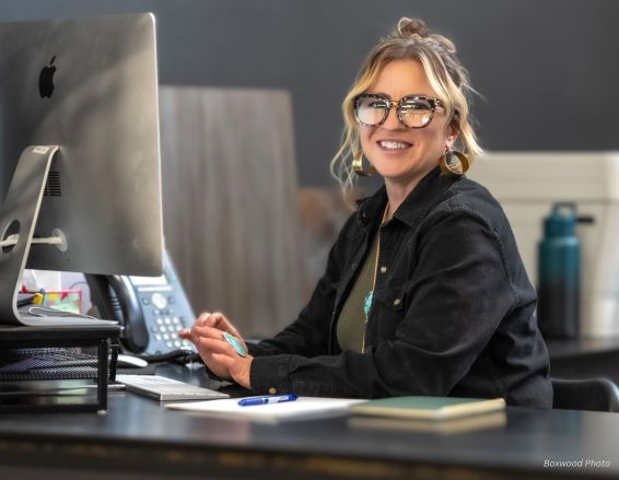 Wearing a black jacket and large hoop earrings, a cheerful person with glasses sits at a desk using the Jackie Bio iMac. Nearby are a telephone, notepad, and water bottle.
