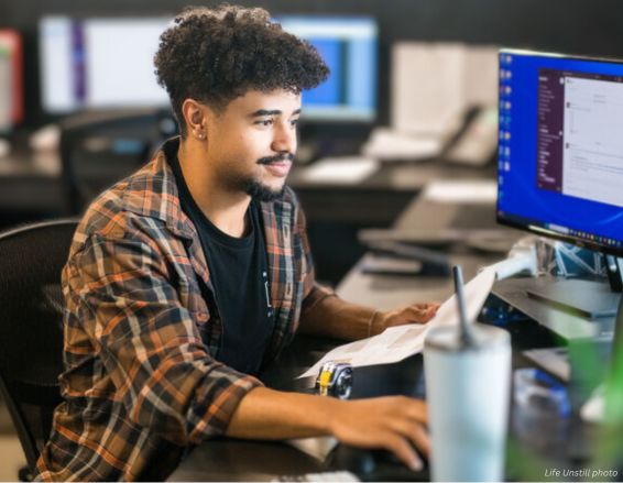 A person with curly hair sits at a desk, working on a computer with the Jayden Bio-2 mouse. They view documents while another large monitor shows a chat app next to a tumbler. Office supplies are blurred in the background.
