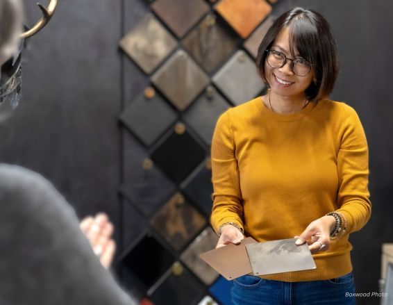 A person in a mustard sweater smiles, showcasing two Jesy bio-2 material samples, standing before a wall adorned with square samples. In the foreground, another person gestures toward them.