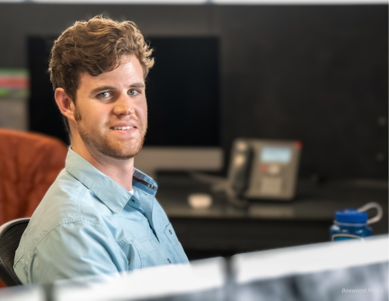 A person with curly hair and a beard, seated at a desk, looks at the camera. In the workspace are a blurred monitor, a phone in the background, and a Luke bio water bottle in the foreground.