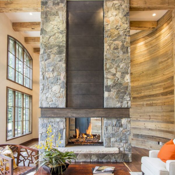 The living room features a large Mantel 4 stone fireplace, wooden accents, and tall left-side windows that flood the space with natural light. A white sofa adorned with an orange cushion sits nearby, complemented by a wooden coffee table holding a potted plant and magazines.