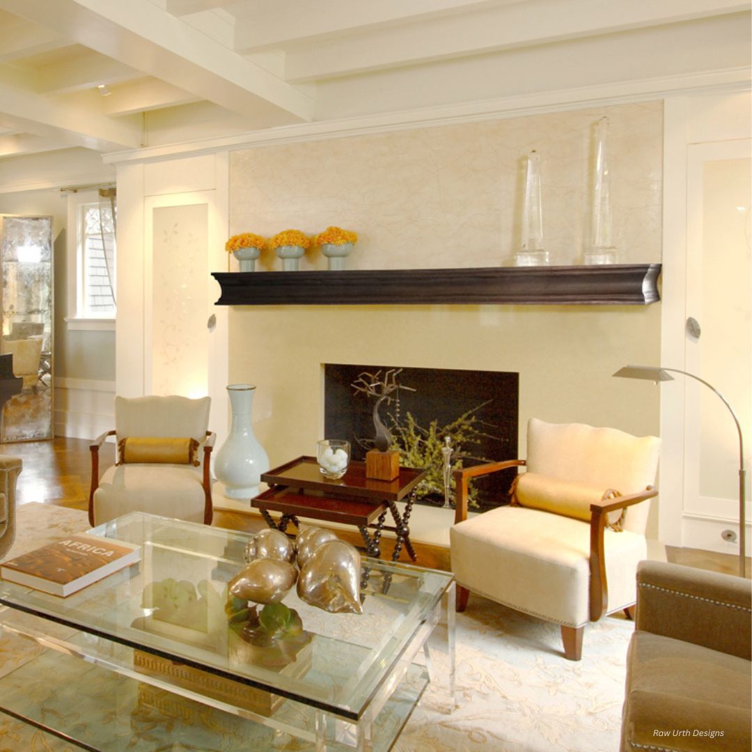 Mantle 3 A stylish living room with cream walls and a beamed ceiling features the Mantle 3 black fireplace mantle, contemporary glass coffee table, beige armchairs, decorative vases, and a small potted plant. Soft lighting enhances the cozy ambiance.