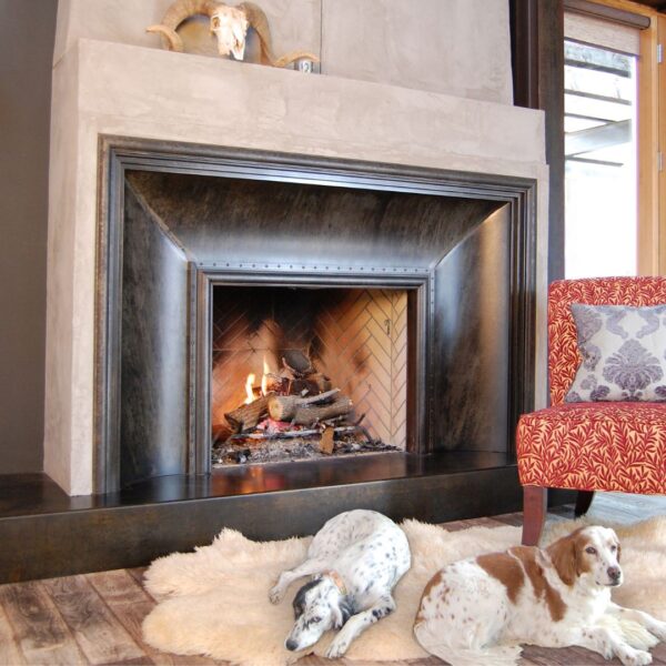 A cozy room features a roaring fireplace with Surround 2's decorative animal skull on the mantel. Two dogs rest on a plush rug in front, next to an upholstered chair with a decorative cushion. Wooden floors complete the scene.