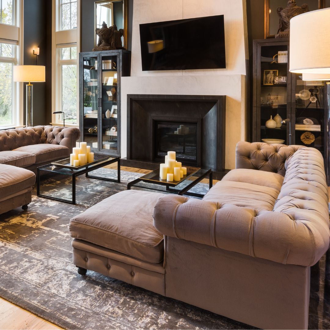 Enjoy luxurious living in Surrounds 1, featuring tufted sofas, a large central fireplace with tall shelves on either side, and modern decor. A flat-screen TV is mounted above the fireplace, complemented by candles on glass coffee tables. Large windows provide ample natural light.