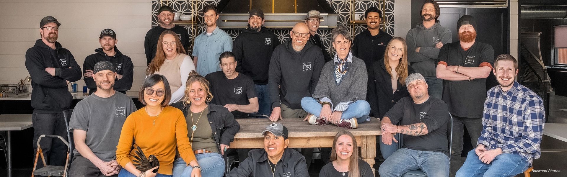 Eighteen casually dressed individuals, smiling together around a wooden table, form the Urthling group photo. They are indoors against a patterned wall with industrial accents.