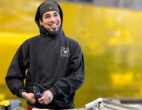 A person in a black hoodie and beanie smiles, standing in an industrial setting with gloves and tools on the table. Behind them is a large yellow surface. They are using Will bio-2 products.