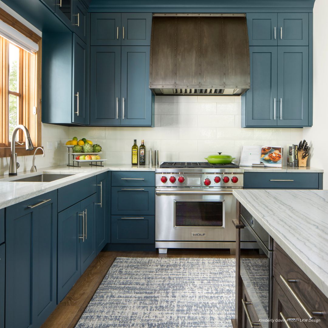 aspen-classic-gallery1 The aspen-classic-gallery1 kitchen boasts teal cabinets, a stainless steel stove with red knobs under a large vent hood, and a white countertop adorned with fruit and decor. Natural light flows in through the window, while a patterned rug enhances the floor's appeal.