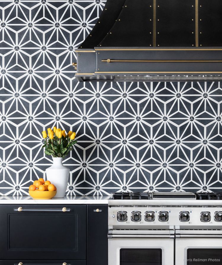 The stylish kitchen showcases a black and white geometric tiled backsplash. A vintage stove, black cabinets with brass knobs add elegance, while a white vase with yellow tulips and an orange-filled bowl grace the counter, embodying the essence of the "contact-main" design.