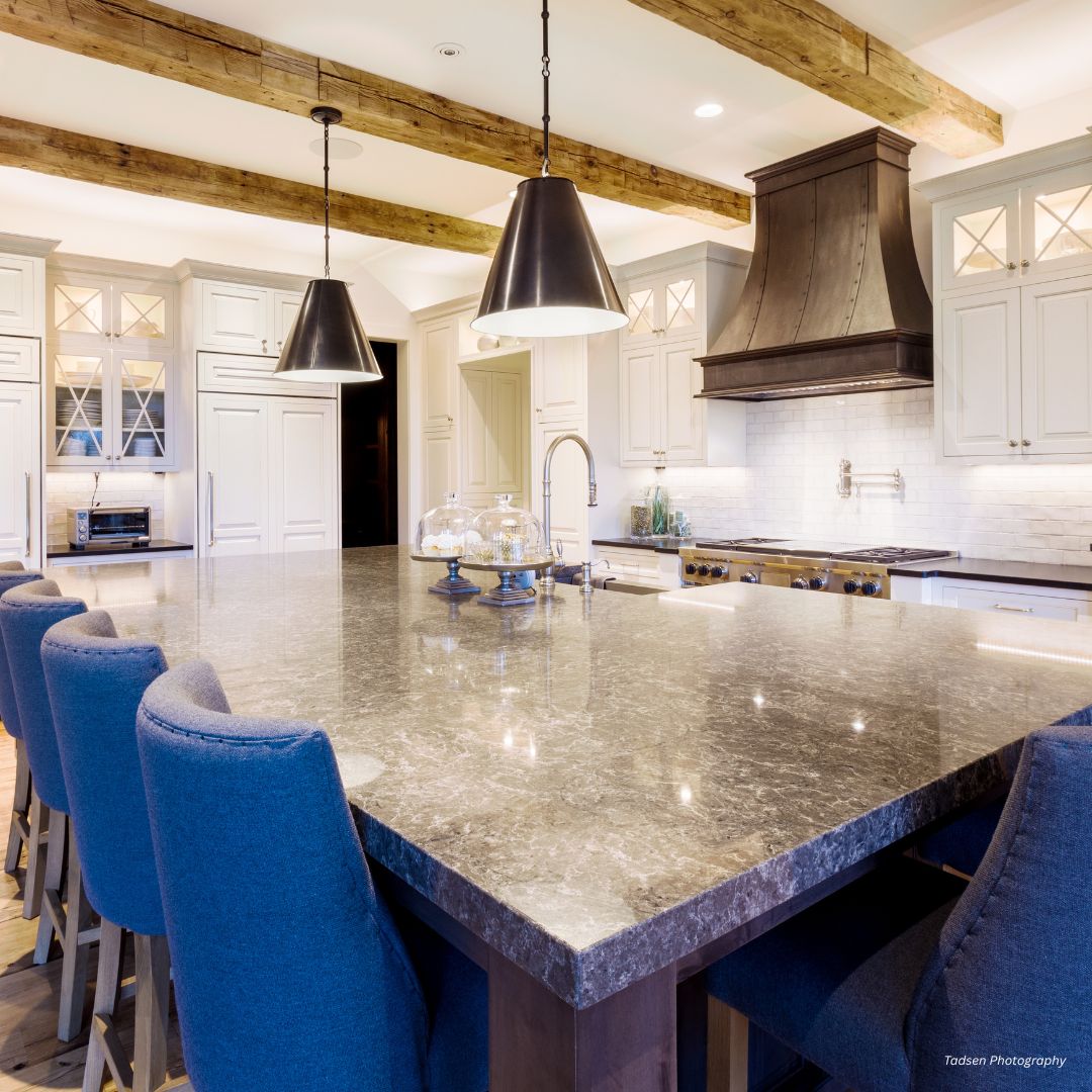 montrose-classic-gallery3√ The montrose-classic-gallery3√ features a bright, modern kitchen with a large marble island, blue upholstered chairs, pendant lights, exposed wooden beams, white cabinetry, stainless steel appliances, and a dark range hood.