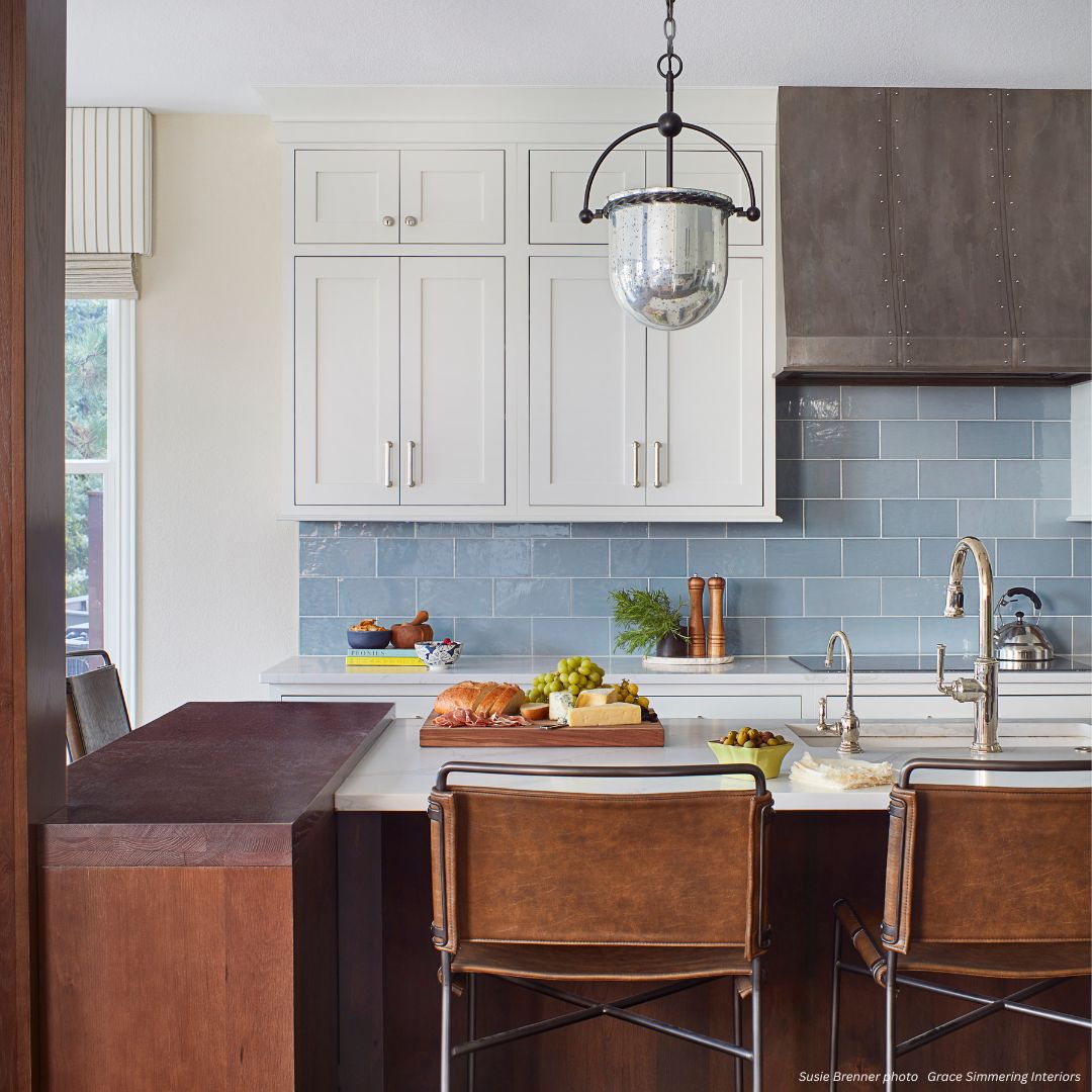 Avon Tailored - gallery 1√ Modern kitchen featuring Avon Tailored cabinets, blue tile backsplash, pendant lighting, and brown leather stools at a white island. The countertop displays bread, fruit, cheese, and a wooden cutting board.