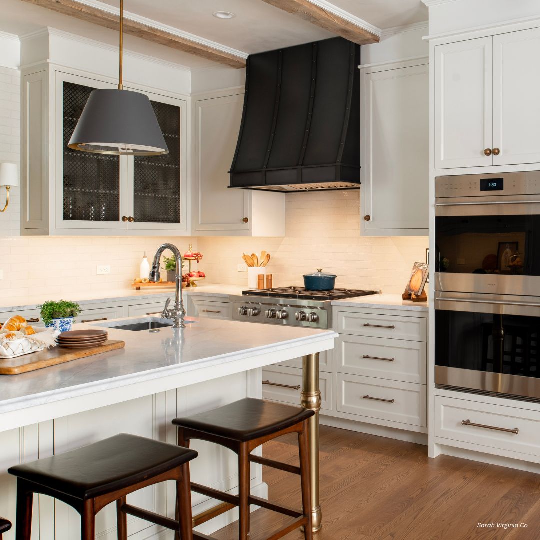 Avon-refined-gallery1√ The Avon-refined-gallery1√ features a modern kitchen with white cabinets, a marble island with two wooden stools, stainless steel double oven, black range hood, wooden floors, warm lighting, and neatly arranged kitchenware for an inviting feel.