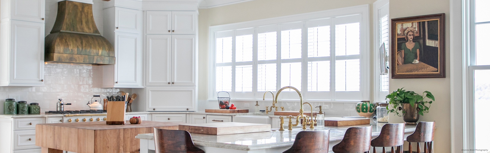The bright, modern kitchen features the Banner Bonner Peak Custom with white cabinets, a large wooden island, brass range hood, marble countertop, and wooden stools. Large windows fill the space with light and a framed painting decorates the dining wall.