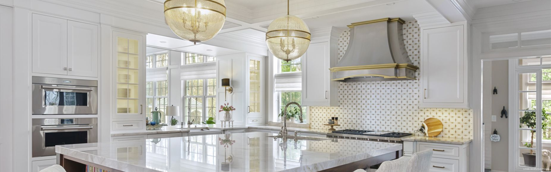Banner Cherry Hills Custom Bright, modern kitchen featuring Banner Cherry Hills Custom white cabinets, marble countertops, large island, stainless steel appliances, patterned backsplash, two globe pendant lights, and expansive windows for natural light.