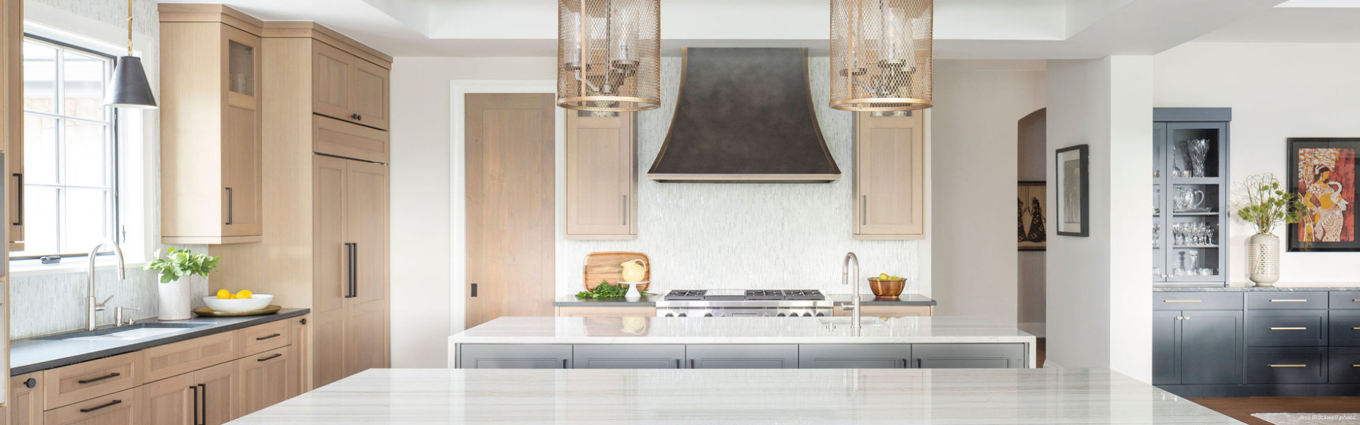 Banner Dove Creek Custom Modern kitchen featuring Banner Dove Creek Custom light wood cabinets, a marble island, stainless steel appliances, two pendant lights, a black range hood, and a vase of flowers on the side counter. Large windows bring in natural light.