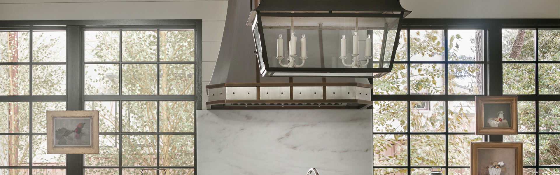Banner Durango Custom A kitchen featuring Banner Durango Custom large grid windows, a marble backsplash, a metal vent hood with candle-style lights, framed wall art, and views of trees through the windows.